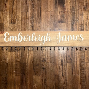 Op de afbeelding: Een houten bord met de namen "Emberleigh James" in witte letters, met een rij zwarte haken eronder.