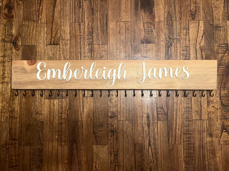 Op de afbeelding: Een houten bord met de namen "Emberleigh James" in witte letters, met een rij zwarte haken eronder.
