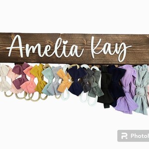 Op de afbeelding: Een houten bord met de naam "Amelia Kay" in witte letters. Het bord heeft haken om spullen aan op te hangen, en er hangen verschillende kleurrijke stoffen haarbanden aan de haken.