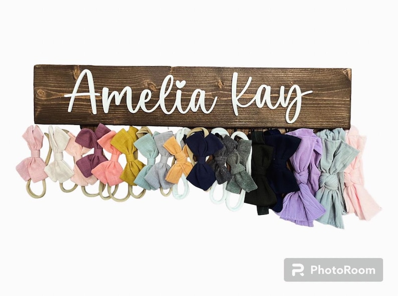 Op de afbeelding: Een houten bord met de naam "Amelia Kay" in witte letters. Het bord heeft haken om spullen aan op te hangen, en er hangen verschillende kleurrijke stoffen haarbanden aan de haken.