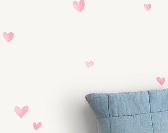 May include: A white wall adorned with scattered, watercolor-style pink hearts. A blue, square pillow is visible in the lower right corner. The hearts vary in size and are evenly distributed across the wall, creating a whimsical and romantic aesthetic.
