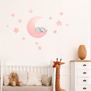 Könnte beinhalten: Ein weißes Babybett mit einer beige Matratze und einem Regenbogen-Teppich. Eine Stoffgiraffe und ein Stoffhase liegen auf dem Babybett. Ein rosa Mond mit einem schlafenden Koala ist an der Wand mit rosa Sternen.