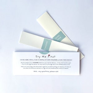 Pode incluir: Duas pequenas amostras brancas de material de decalque com etiquetas que dizem "Standard" e "High Tack". As amostras estão sobre uma superfície branca com um cartão azul e branco que diz "Try Me First" e explica a diferença entre os dois tipos de material de decalque.