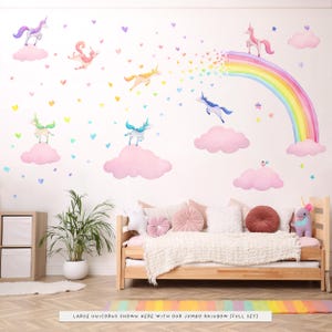 Op de afbeelding: Een grillige muursticker set met aquarel eenhoorns, wolken, een regenboog en harten in pastelkleuren. De sc&egrave;ne omvat een bed met kussens en een tapijt met een regenboogpatroon. De tekst onderaan luidt "LARGE UNICORNS SHOWN HERE WITH OUR JUMBO RAINBOW (FULL SET)".