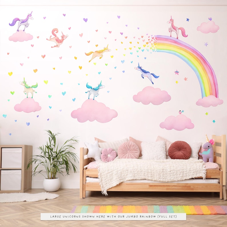 Op de afbeelding: Een grillige muursticker set met aquarel eenhoorns, wolken, een regenboog en harten in pastelkleuren. De sc&egrave;ne omvat een bed met kussens en een tapijt met een regenboogpatroon. De tekst onderaan luidt "LARGE UNICORNS SHOWN HERE WITH OUR JUMBO RAINBOW (FULL SET)".