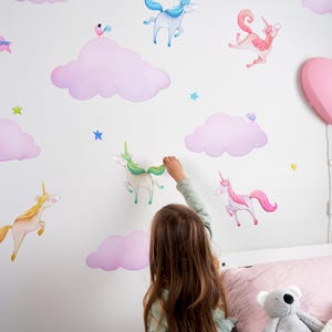 Op de afbeelding: Een kind versiert een witte muur met aquarel eenhoorn- en wolkenstickers. De stickers tonen eenhoorns in verschillende kleuren, waaronder roze, geel en blauw, met bijpassende roze wolken en blauwe sterren.