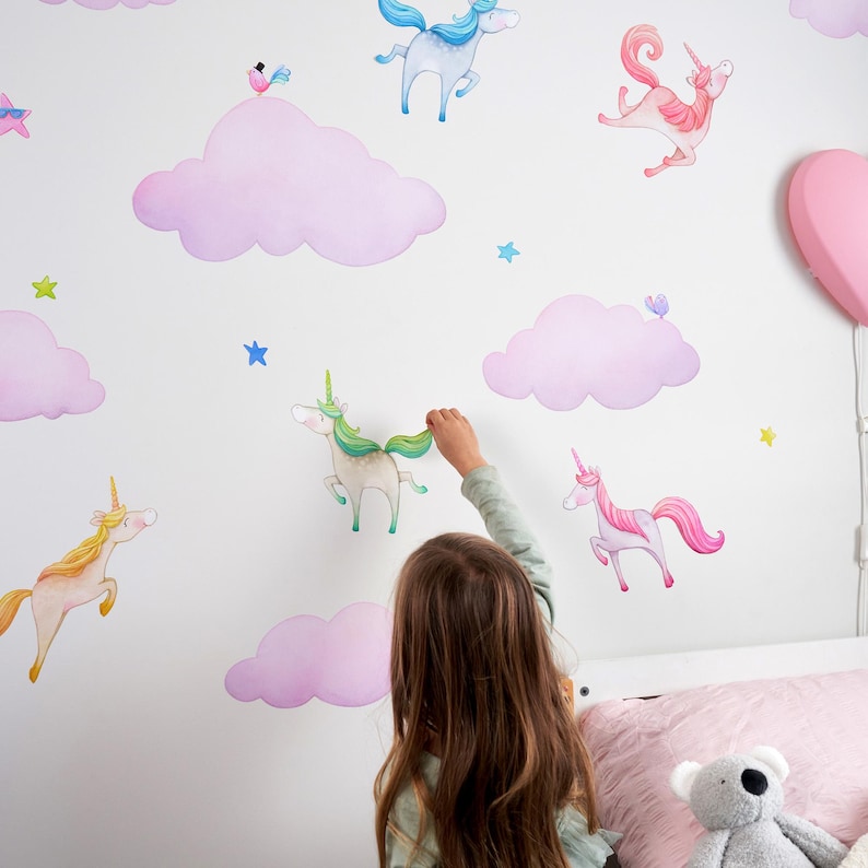 Op de afbeelding: Een kind versiert een witte muur met aquarel eenhoorn- en wolkenstickers. De stickers tonen eenhoorns in verschillende kleuren, waaronder roze, geel en blauw, met bijpassende roze wolken en blauwe sterren.
