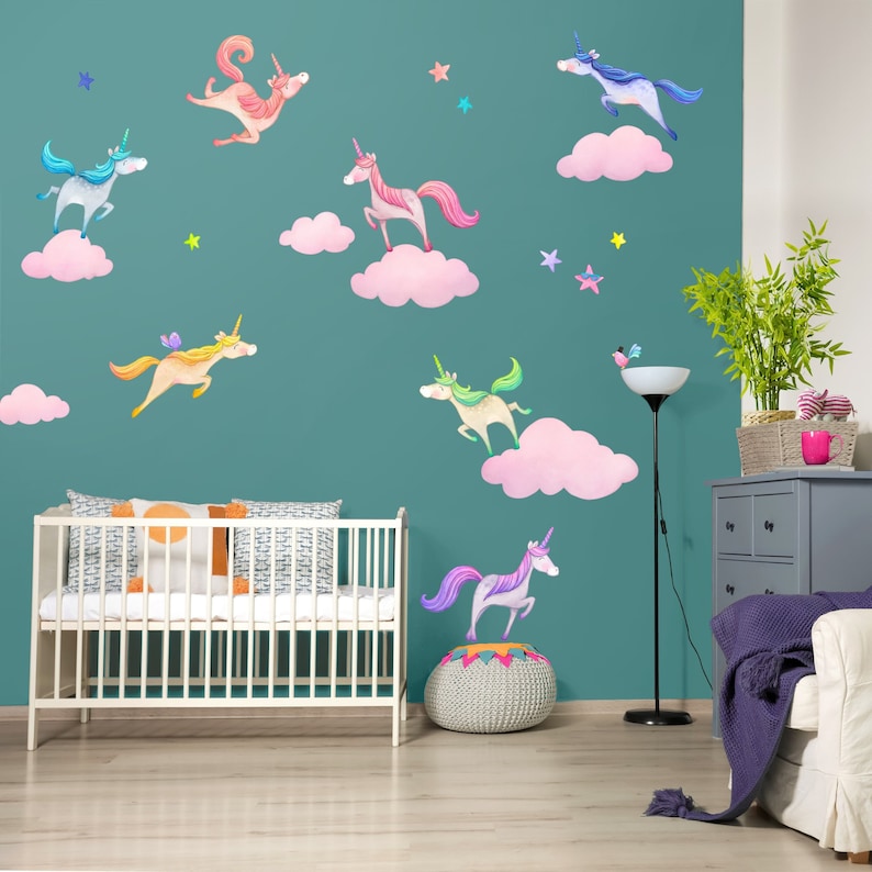 Op de afbeelding: Een turquoise muur versierd met aquarel eenhoornstickers, roze wolken en sterren. Op de voorgrond staan een witte wieg met patronen kussens en een grijze ladekast. Een vloerlamp en een witte bank zijn ook zichtbaar.