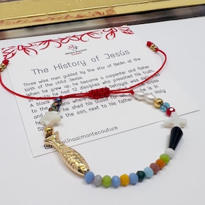 Puede incluir: Un brazalete de cuerda roja con un dije de pez dorado, cuentas blancas y negras y un dije de cruz blanca. El brazalete está sobre una tarjeta blanca con el texto "The History of Jesús" y una descripción de la vida de Jesús.