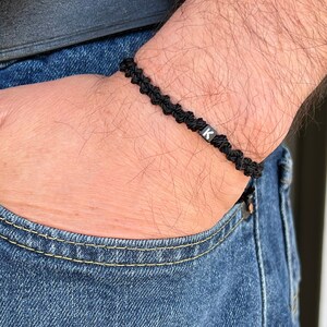 Pode incluir: Um bracelete trançado preto com um fecho de letra 'K' preto. O bracelete é usado no pulso.