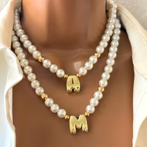 Puede incluir: Collar de perlas de dos hilos con detalles dorados y colgantes de letras doradas. El collar superior presenta la letra "A", y el collar inferior presenta la letra "M". Las perlas son blancas y las letras son doradas.