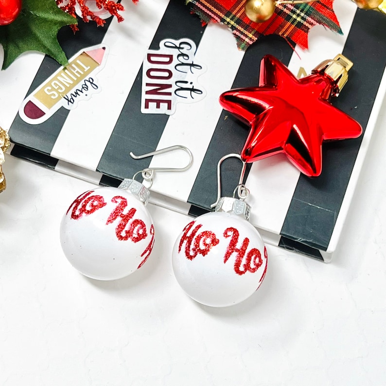 May include: A pair of silver earrings with white ornament-shaped charms that say "Ho Ho Ho" in red glitter lettering.