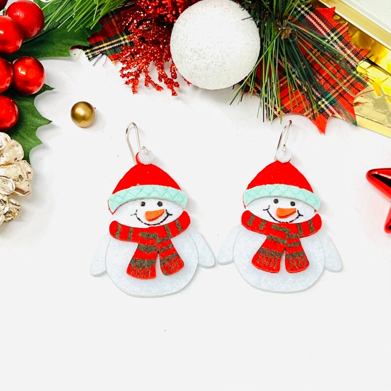May include: A pair of snowman-shaped earrings with red hats, green trim, and red and brown striped scarves. The earrings are made of felt and have silver hooks.