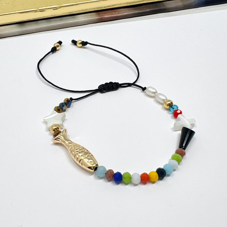 May include: A colorful beaded bracelet with a gold fish charm, white shell charms, and a black and white shell charm. The bracelet is adjustable with a black cord.