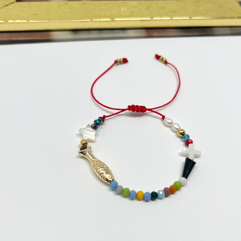 May include: A red string bracelet with colorful beads, a gold fish charm, a white star charm, and a white cross charm.