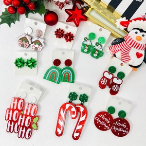 May include: A collection of Christmas-themed earrings in various designs. Styles include gingerbread houses, bows, mugs with snowmen, mittens with snowflakes, "HO HO HO" text, candy canes, and "Merry Christmas" circles. The earrings are red, green, and white.