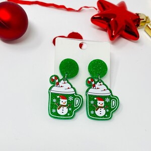 May include: A pair of green glitter Christmas-themed earrings featuring a snowman in a mug with whipped cream and candy canes.