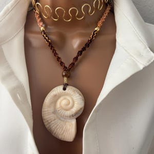 Puede incluir: Un collar de cordón trenzado marrón y beige con un collar gargantilla de cadena dorada y un gran colgante de concha blanca. El colgante de concha está tallado para parecerse a un nautilus.