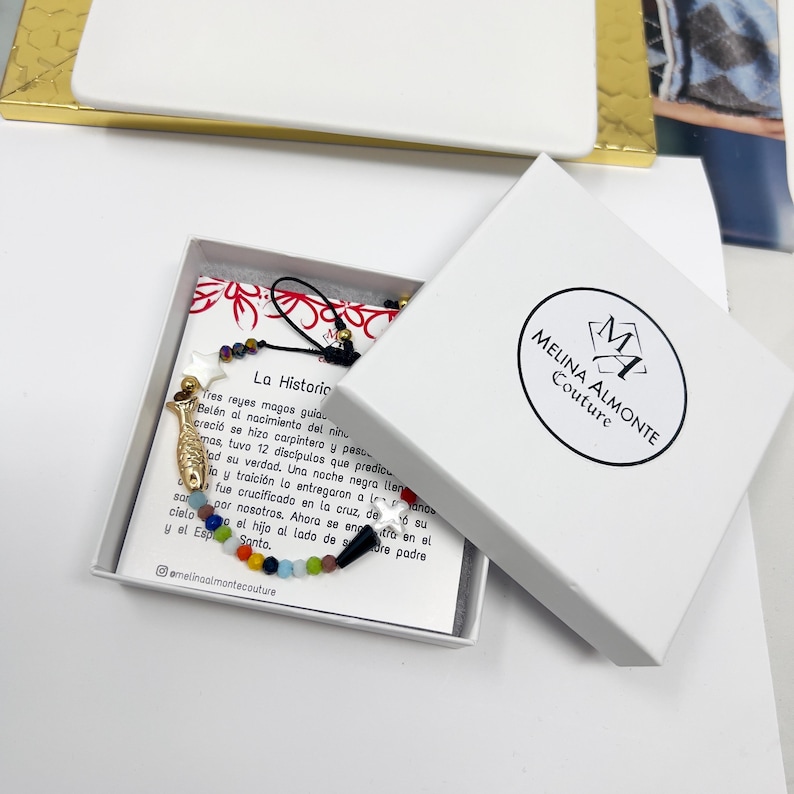 May include: A white box with a black and red logo that says "Melina Almonte Couture". The box is open and contains a colorful beaded necklace with a gold fish charm and a black and white charm. The necklace is on a white card with the text "La Historic" and a description of Jesus Christ in Spanish.