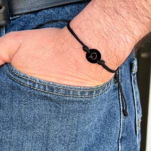 Peut inclure: Un bracelet en cordon noir avec un charme circulaire noir représentant une lettre stylisée "G".
