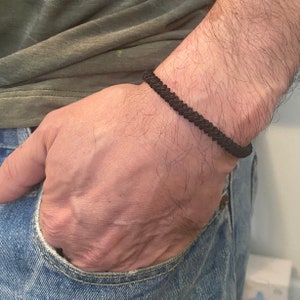 May include: A black braided bracelet is worn on a wrist. The bracelet is made of a series of tightly woven strands, creating a textured appearance. The bracelet is a simple, yet stylish accessory.