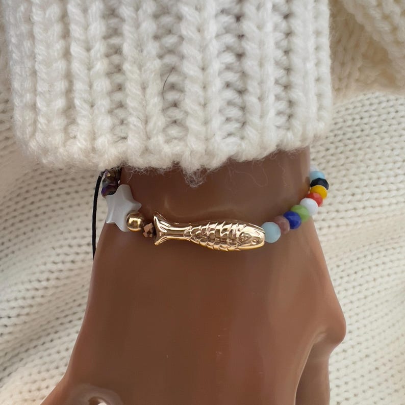 May include: A colorful beaded bracelet with a gold fish charm. The bracelet is made with a variety of colored beads, including blue, green, red, and yellow. The fish charm is attached to the bracelet with a black cord.