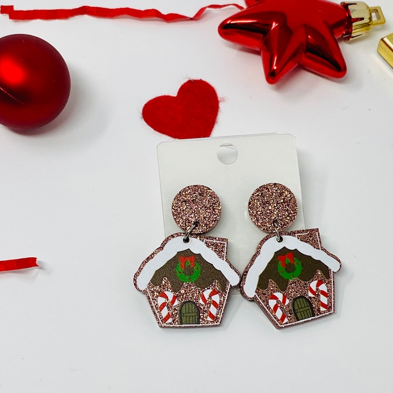May include: A pair of gingerbread house earrings with glitter accents. The earrings are brown, white, and green with red accents. The earrings are on a white background.