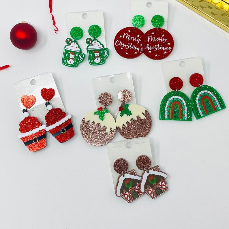 May include: Assortment of Christmas-themed earrings featuring various designs, including a gingerbread house, a snowman, a pudding, a cupcake, and a rainbow. The earrings are made of glitter and have a festive red and green color scheme.