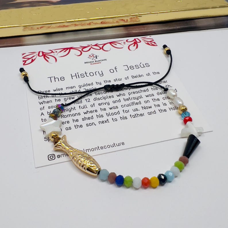 May include: A colorful beaded bracelet with a gold fish charm, a white shell, and a black and white cross charm. The bracelet is on a card with the text "The History of Jes&uacute;s" and a quote about the story of Jesus.