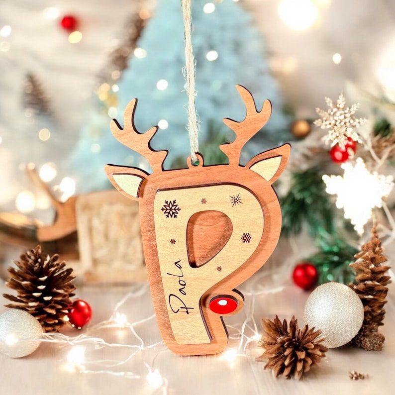 May include: Wooden Christmas ornament shaped like a reindeer head with the letter "P" cut out. The ornament is decorated with snowflakes and the name "Paola" is engraved on the side.