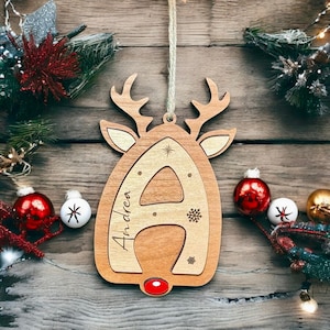 May include: Wooden reindeer ornament with the letter A and the name Andrea engraved on it. The ornament is decorated with snowflakes and stars. It has a red nose and a string for hanging.