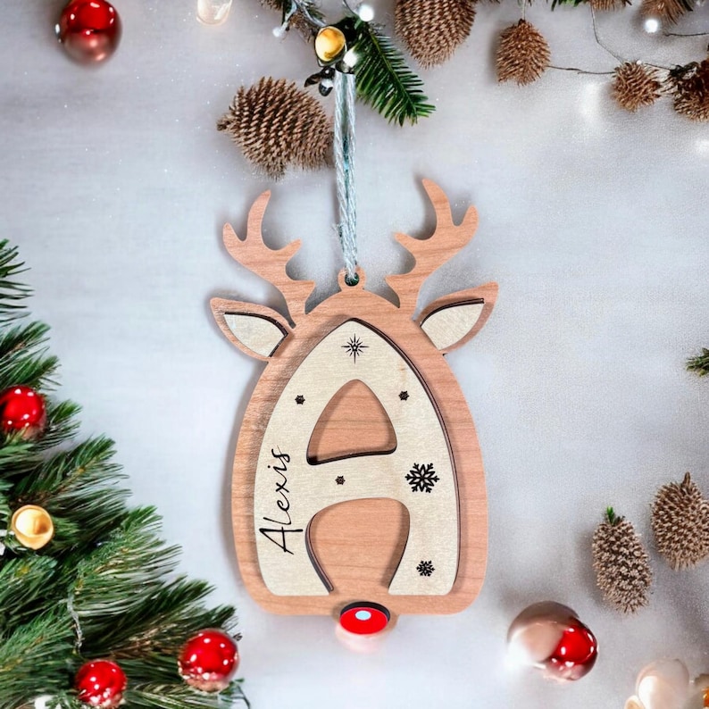 May include: Wooden Christmas ornament shaped like a reindeer with the letter A cut out and the name Alexis written on it. The ornament is decorated with snowflakes.