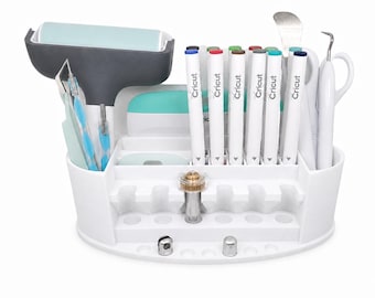 Puede incluir: Un organizador de herramientas de manualidades de plástico blanco que contiene varios suministros, incluyendo un rodillo, seis marcadores de la marca Cricut, una espátula de metal, una herramienta de depilado y varios accesorios metálicos.