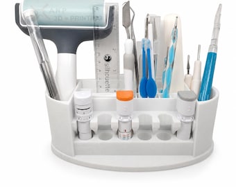 Puede incluir: Un organizador de herramientas de manualidades de plástico blanco que contiene varios utensilios, incluyendo un rodillo, una regla transparente con marcas de 3 pulgadas, espátulas de metal, punzones azules y blancos y tres cuchillas cilíndricas.