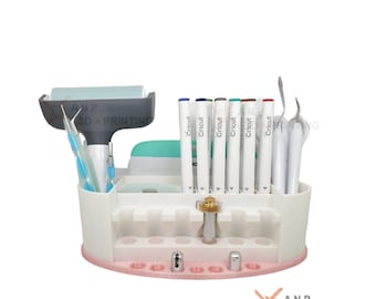 Puede incluir: Organizador blanco con ribete rosa para guardar artículos de artesanía. Tiene ranuras para bolígrafos, marcadores y herramientas. El organizador está lleno de bolígrafos y marcadores de la marca Cricut, una herramienta de raspado y una herramienta de deshierbe.