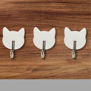 May include: Three white cat-shaped hooks with silver metal hooks. The hooks are mounted on a wood surface.