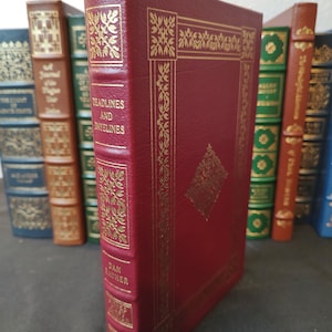 Pode incluir: Um livro encadernado em couro vermelho com detalhes dourados e o título "Deadlines and Datelines" de Dan Rather.