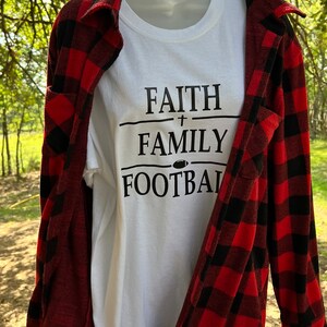 May include: White t-shirt with black text that reads "FAITH + FAMILY FOOTBALL". The shirt is being worn by a mannequin and is layered over a red and black plaid shirt.