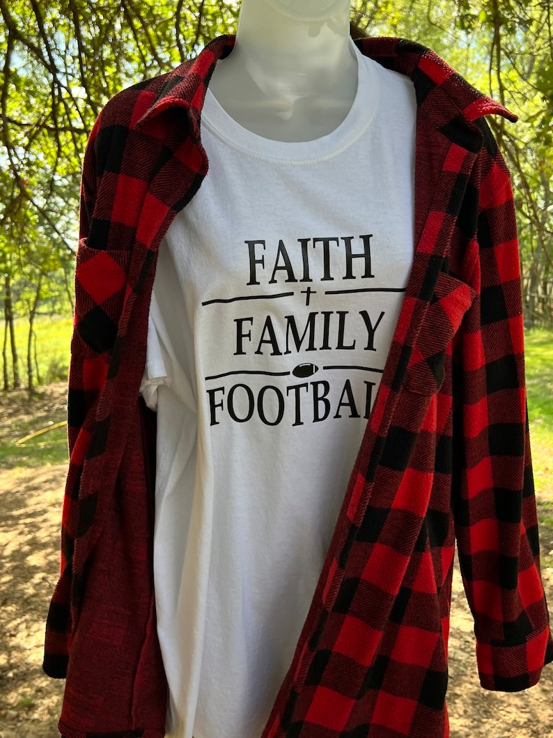 May include: White t-shirt with black text that reads "FAITH + FAMILY FOOTBALL". The shirt is being worn by a mannequin and is layered over a red and black plaid shirt.