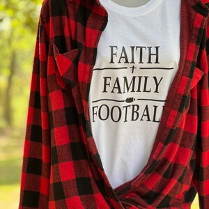 May include: White t-shirt with black text that reads "Faith + Family Football". The shirt is layered over a red and black checked flannel shirt.