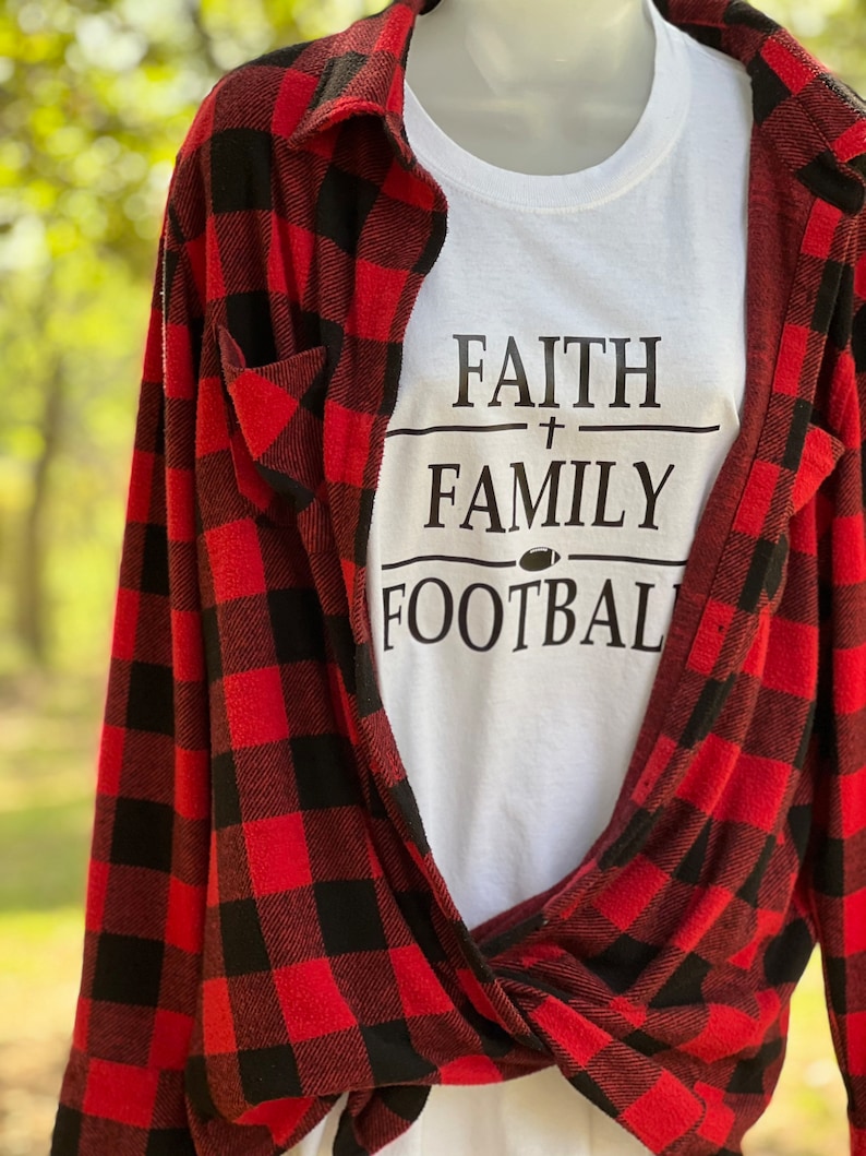 May include: White t-shirt with black text that reads "Faith + Family Football". The shirt is layered over a red and black checked flannel shirt.