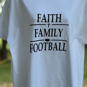May include: White t-shirt with black text that reads "FAITH + FAMILY FOOTBALL" with a football icon between the words "FAMILY" and "FOOTBALL".
