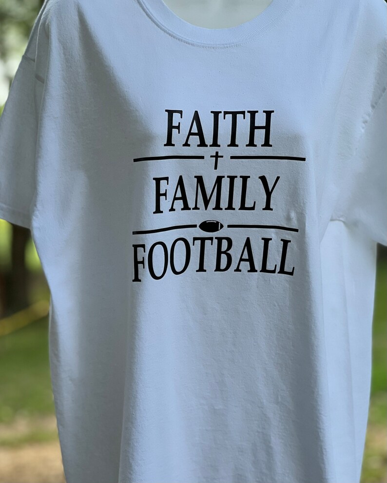 May include: White t-shirt with black text that reads "FAITH + FAMILY FOOTBALL" with a football icon between the words "FAMILY" and "FOOTBALL".