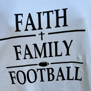 May include: White t-shirt with black text that reads "FAITH + FAMILY FOOTBALL" with a football graphic between the words "FAMILY" and "FOOTBALL".