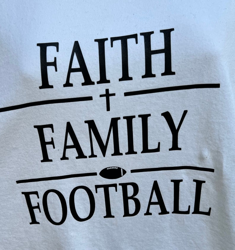 May include: White t-shirt with black text that reads "FAITH + FAMILY FOOTBALL" with a football graphic between the words "FAMILY" and "FOOTBALL".
