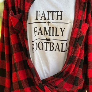 May include: White t-shirt with black text that reads "FAITH + FAMILY FOOTBALL" with a football graphic. The shirt is layered over a red and black checked flannel shirt.