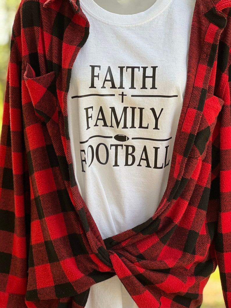 May include: White t-shirt with black text that reads "FAITH + FAMILY FOOTBALL" with a football graphic. The shirt is layered over a red and black checked flannel shirt.