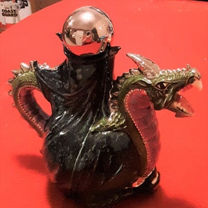 Può includere: Una teiera a forma di drago verde e argento con un coperchio a sfera argentato. Il drago ha una bocca spalancata con denti affilati e una lingua rosa.