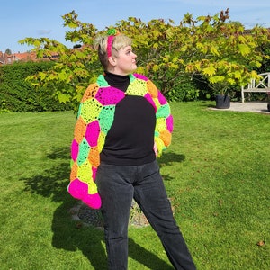 Puede incluir: Un chaleco de crochet hecho a mano con un patrón geométrico en rosa neón, naranja y verde. El chaleco se lleva sobre un cuello alto negro y se combina con vaqueros negros. Una diadema a juego completa el look, sobre un fondo soleado al aire libre.