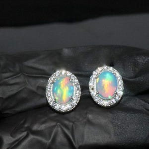 May include: A pair of silver earrings featuring oval opal gemstones set in a halo of smaller clear stones.